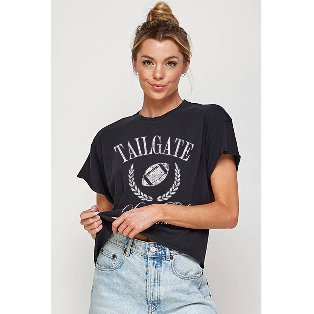 Studio portrait photography of a woman in a white studio wearing a distressed black t-shirt featuring white TAILGATE text and a laurel-framed football graphic paired with light-wash denim jeans and a high hair bun