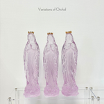 Three translucent orchid colored glass statues of the Virgin Mary with gold crowns on a clear acrylic shelf