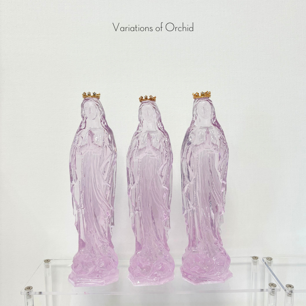 Three translucent orchid colored glass statues of the Virgin Mary with gold crowns on a clear acrylic shelf