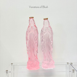 Two translucent pink resin statues of the Virgin Mary with small gold crowns on a clear acrylic shelf