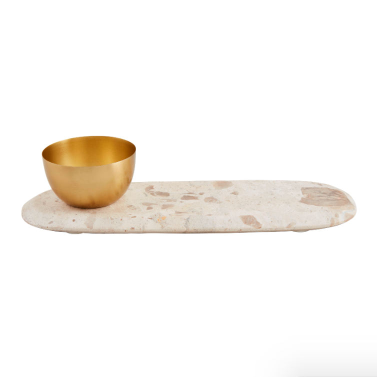 A modern decorative serving set featuring an oval light speckled travertine tray paired with a small shiny gold metal bowl