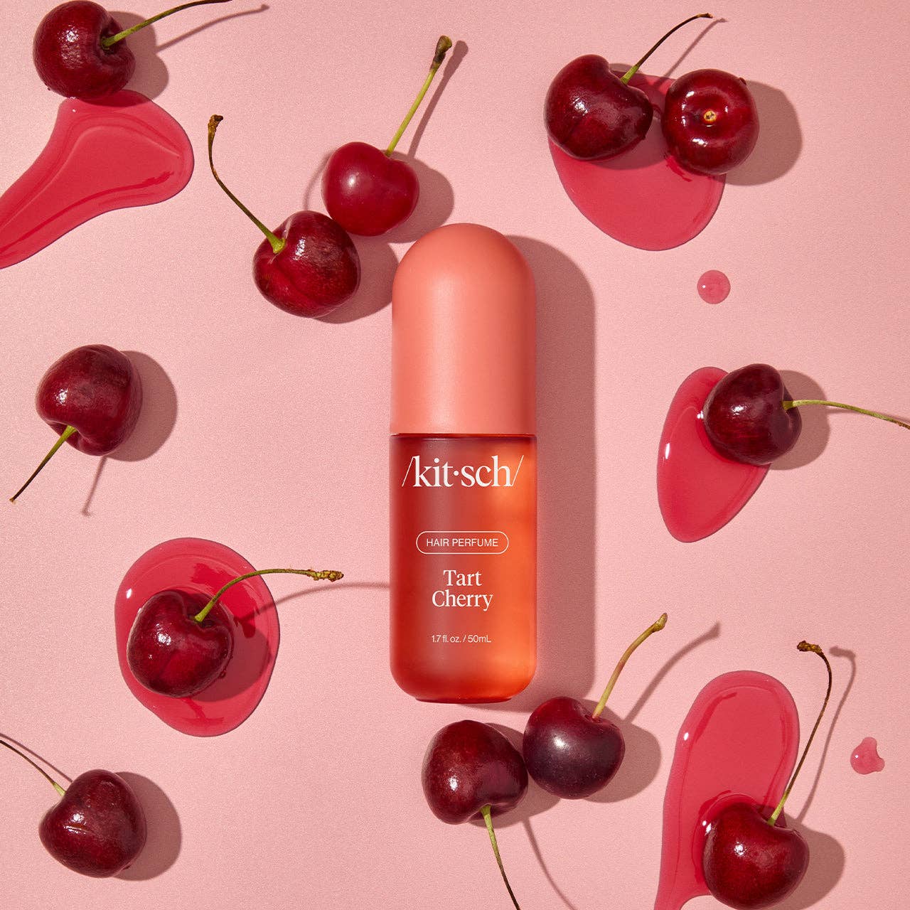Kitsch Tart Cherry hair perfume bottle surrounded by fresh cherries and pink liquid splashes on a pink background