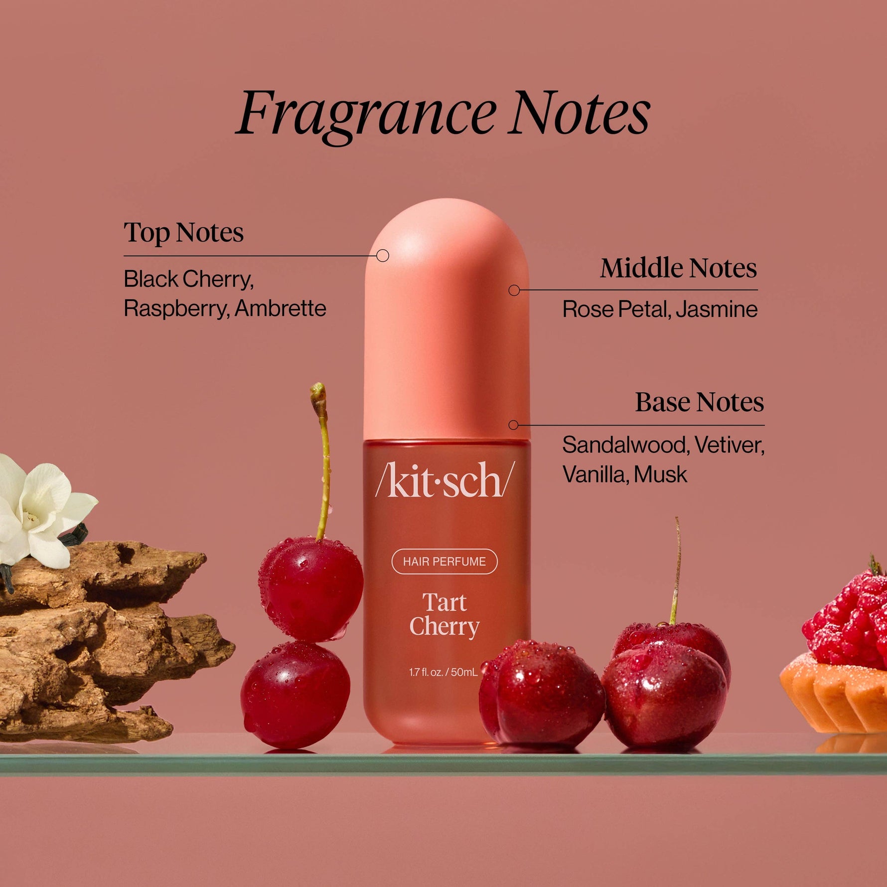 Product photograph of Kitsch Tart Cherry Hair Perfume, a pink bottle displayed on a glass shelf with fresh cherries, a white flower and bark, and a raspberry tart, detailing fragrance notes including black cherry rose petal and sandalwood on a soft pink background