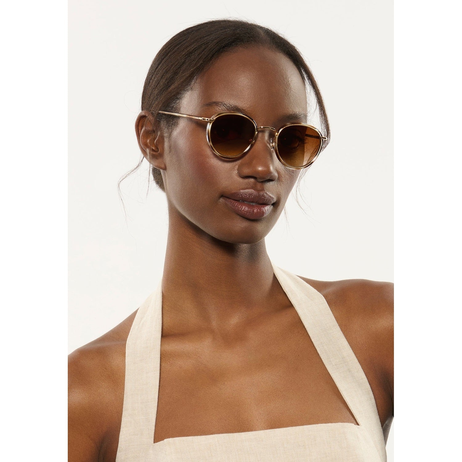 A close-up portrait of a dark-skinned woman with pulled-back hair wearing round gold-rimmed sunglasses with amber gradient lenses and a cream linen halter-neck top against a white studio background