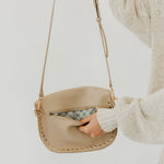 A hand opens a light beige pebbled crossbody bag with gold studded embellishments along the bottom edge and a patterned interior lining worn by a person in a cream knit sweater