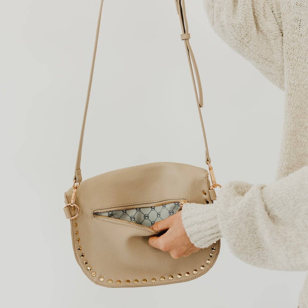 A hand opens a light beige pebbled crossbody bag with gold studded embellishments along the bottom edge and a patterned interior lining worn by a person in a cream knit sweater