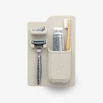 Studio product photograph of a minimalist stone-grey silicone shower organizer holding an iconic Marvis toothpaste tube, a polished chrome razor, and a natural wooden toothbrush covered in decorative water droplets