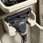 Photograph of a wet modern razor resting in a beige plastic shower organizer, featuring intricate water droplets on a gray textured grip and a multi-blade head suspended in a ribbed wall-mounted holder
