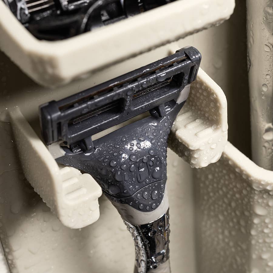 Photograph of a wet modern razor resting in a beige plastic shower organizer, featuring intricate water droplets on a gray textured grip and a multi-blade head suspended in a ribbed wall-mounted holder