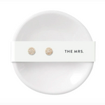 Product photograph showcasing a pair of round pavé diamond gold stud earrings on a white card with THE MRS text, resting in a minimalist white ceramic ring dish
