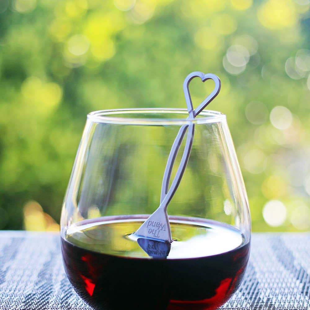 Photograph of a clear stemless wine glass filled with dark red wine featuring a silver heart-shaped The Wand wine purifier immersed on a textured grey outdoor table against a vibrant blurred green foliage background