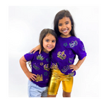 Photograph of two smiling young girls in a studio wearing purple t-shirts adorned with sequined gold, green, and purple Mardi Gras crowns and masks, one girl in shiny gold bike shorts and the other in light blue denim shorts