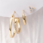 Gold jewelry display featuring a double-layered hoop earring with a row of clear stones, a small plain gold hoop, and a three-stone triangular stud earring on a white background