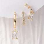 A close-up of three gold-plated earrings on a white background, featuring a hoop with a dangling rectangular cubic zirconia, a pave-set mini hoop, and a curved ear climber with three baguette-cut cubic zirconia