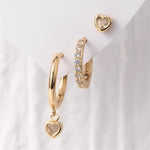 Jewelry presenting three gold earrings: a smooth hoop with a heart charm, a pavé gemstone hoop, and a heart stud, displayed on a clean white surface