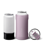 Product photograph of a white beverage can in a black holder next to a dusty lavender Brumate insulated can cooler holding a white can, with a clear flip-top lid featuring a lavender clasp on a white background