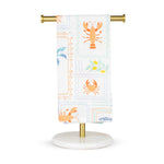 White cotton kitchen towel on a brass and white marble stand featuring a coastal pattern of orange lobsters, orange crabs, blue palm trees, and olive branches with yellow olives in postage stamp-like frames