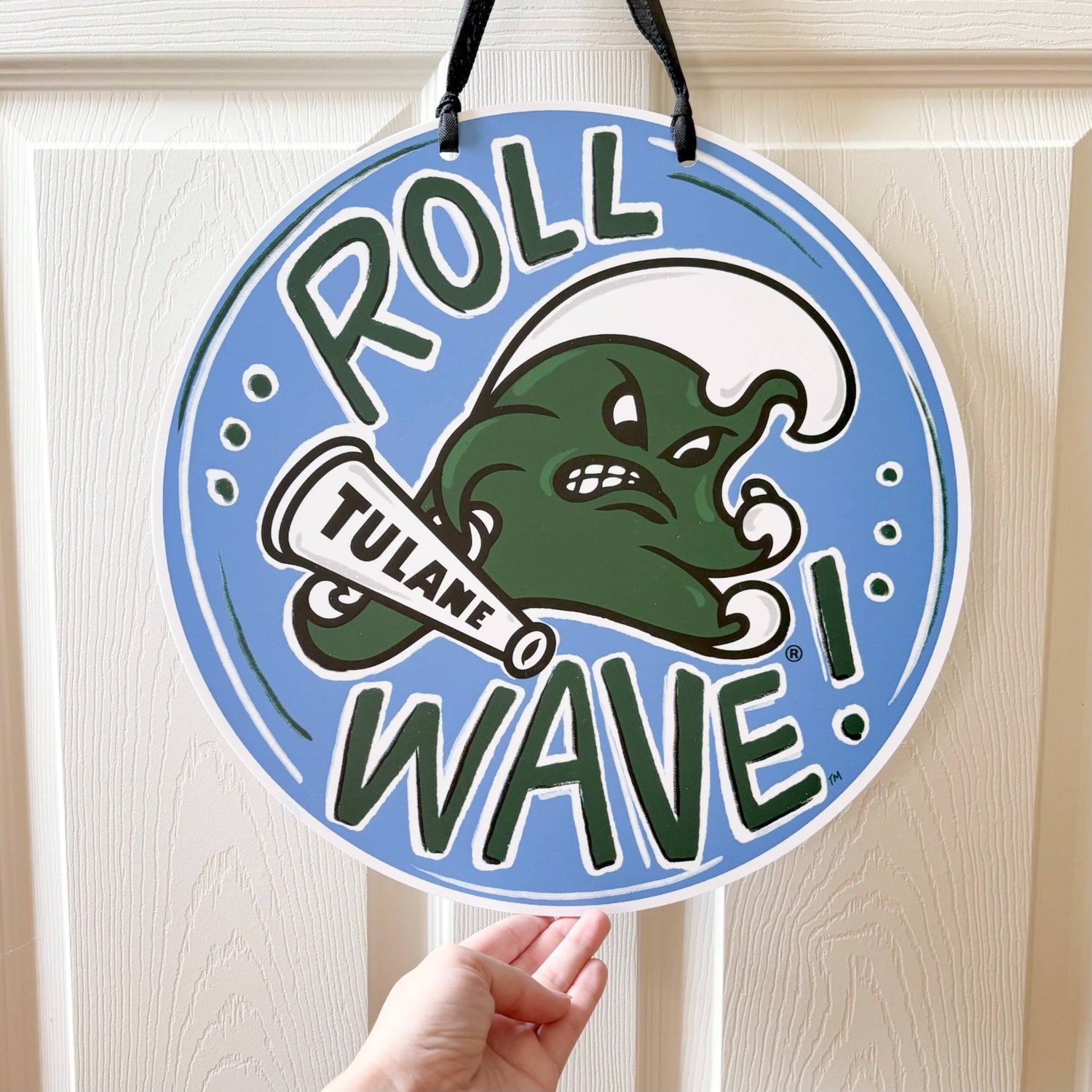 Printed circular sign showing a blue and green Tulane University 'Roll Wave!' design with an angry green wave mascot, a megaphone reading 'Tulane', held by a hand against a white door