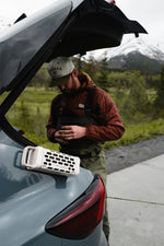 Photograph of a man in a Seager cap and red puffer jacket over waders adjusting gear near a teal car with a white Turtlebox speaker magnetically attached, set against a backdrop of green trees and a snow-capped mountain
