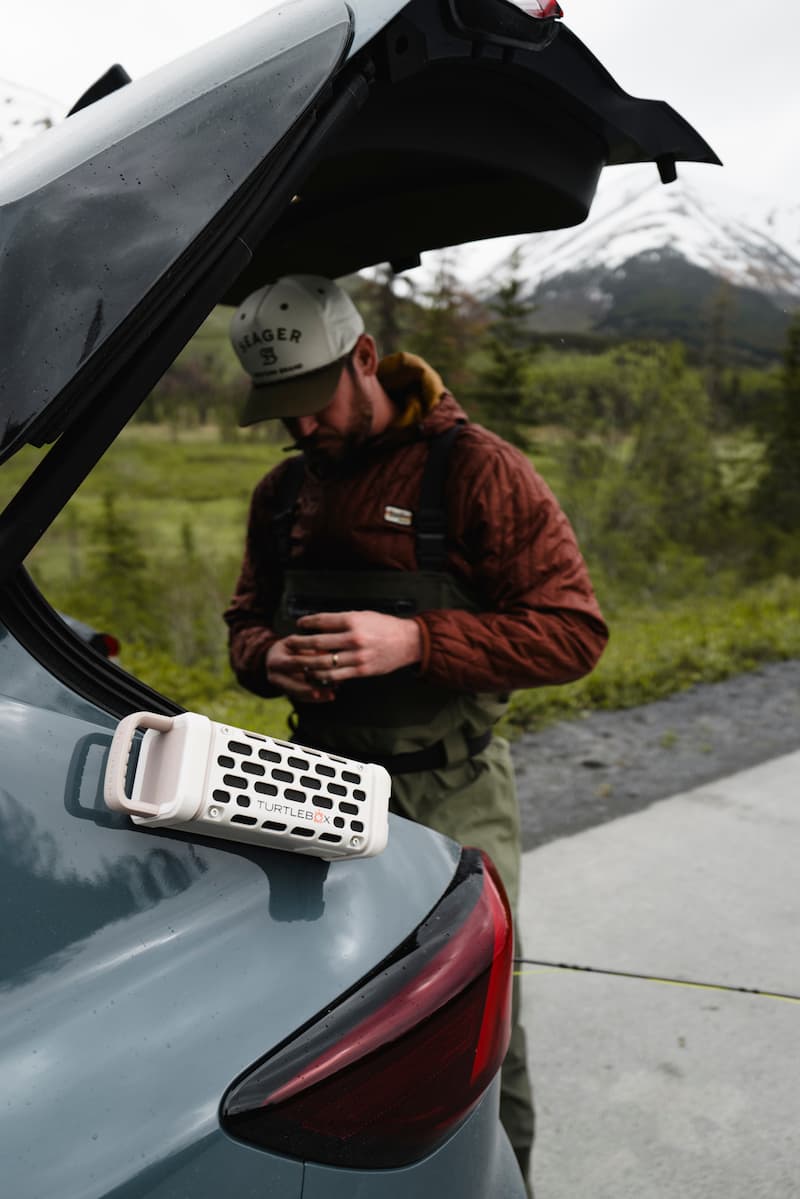 Photograph of a man in a Seager cap and red puffer jacket over waders adjusting gear near a teal car with a white Turtlebox speaker magnetically attached, set against a backdrop of green trees and a snow-capped mountain