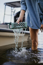 A person wearing a blue striped shirt stands in dark water holding a green Turtlebox speaker with water dripping and splashing from it next to a boat in the background