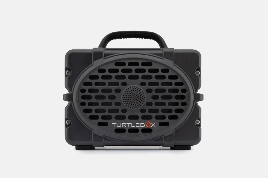 Product photography of a dark grey Turtlebox Gen 2 portable speaker with a rugged design a prominent oval speaker grille a central circular tweeter a top carry handle and a white TURTLEBOX logo with an orange X on a plain white studio background