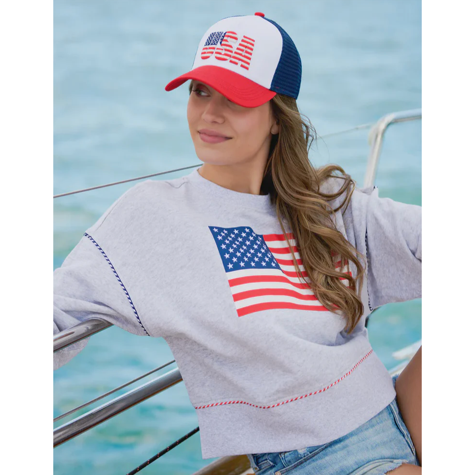Woman wearing a grey sweatshirt with an American flag print and a red white and blue USA trucker hat on a boat