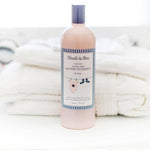 Light pink Noodle & Boo Ultra-Safe Laundry Detergent for baby bottle with lavender cap and a striped label showing a star onesie and blue booties, placed before a stack of white towels