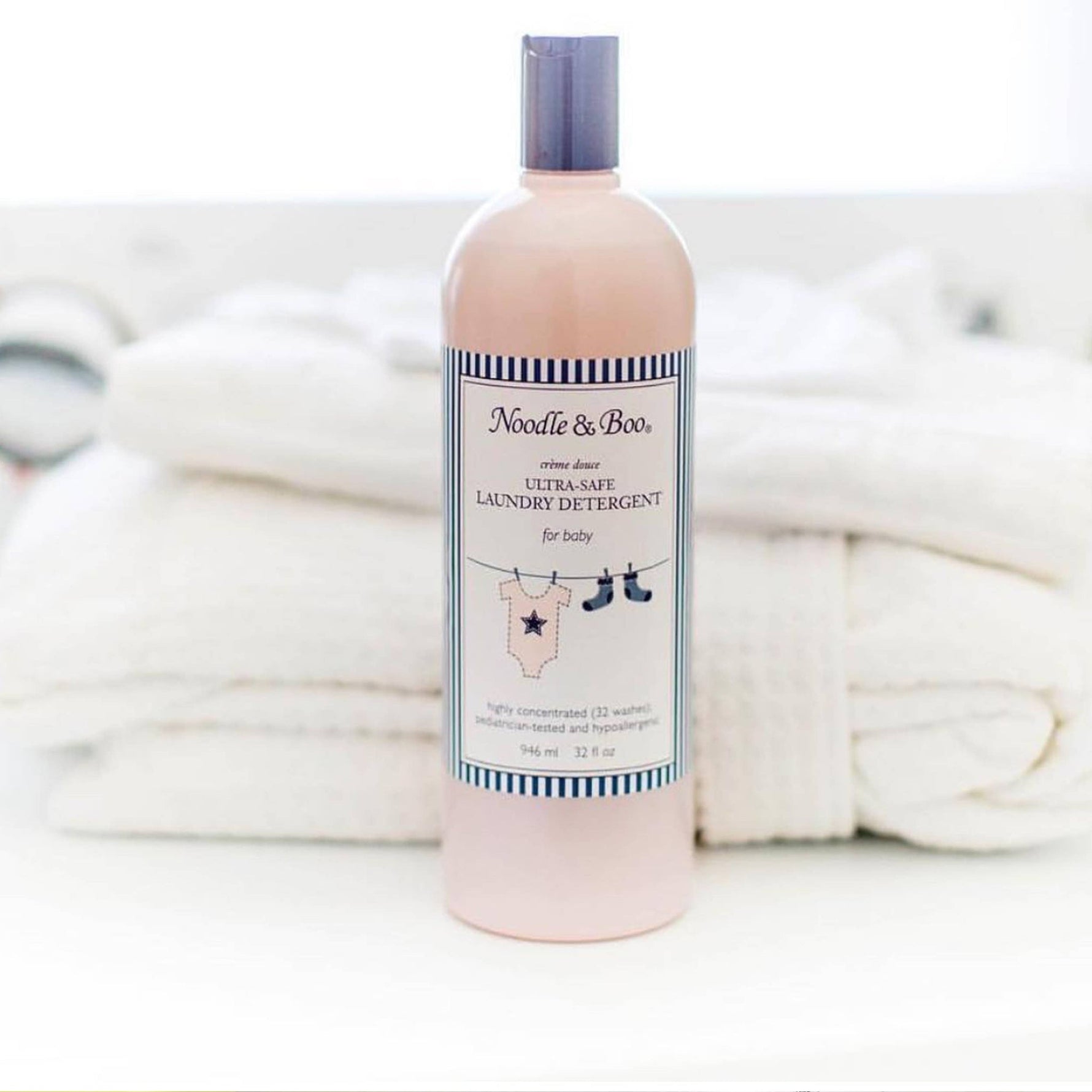 Light pink Noodle & Boo Ultra-Safe Laundry Detergent for baby bottle with lavender cap and a striped label showing a star onesie and blue booties, placed before a stack of white towels