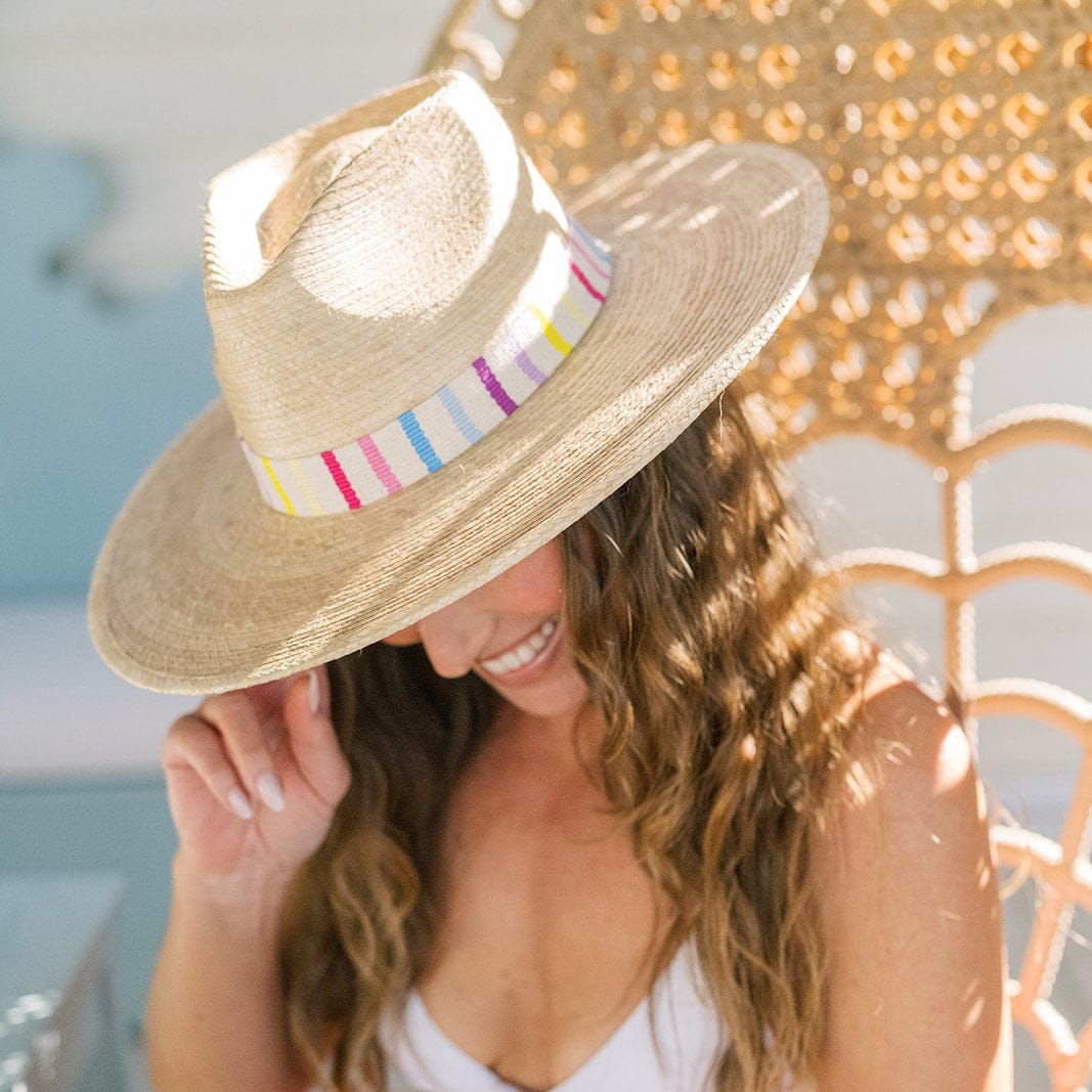 Photograph of a smiling woman with long wavy brown hair, wearing a light straw fedora hat with a vibrant pink, yellow, blue, and purple striped band, relaxing outdoors in a woven rattan chair