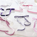 Photography of multiple clear wine glasses adorned with velvet ribbons in dusty rose, rich purple, and soft grey tied around their stems on a clean, reflective white surface