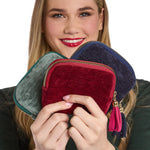 Digital studio photograph of a smiling blonde woman holding three plush velvet zippered pouches in jewel tones of emerald, burgundy, and navy blue accented by gold hardware and vibrant decorative tassels