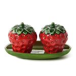 Ceramic pair of strawberry-shaped salt and pepper shakers featuring vibrant red bodies, dark green leafy tops, and a glossy finish resting on a matching green oval tray