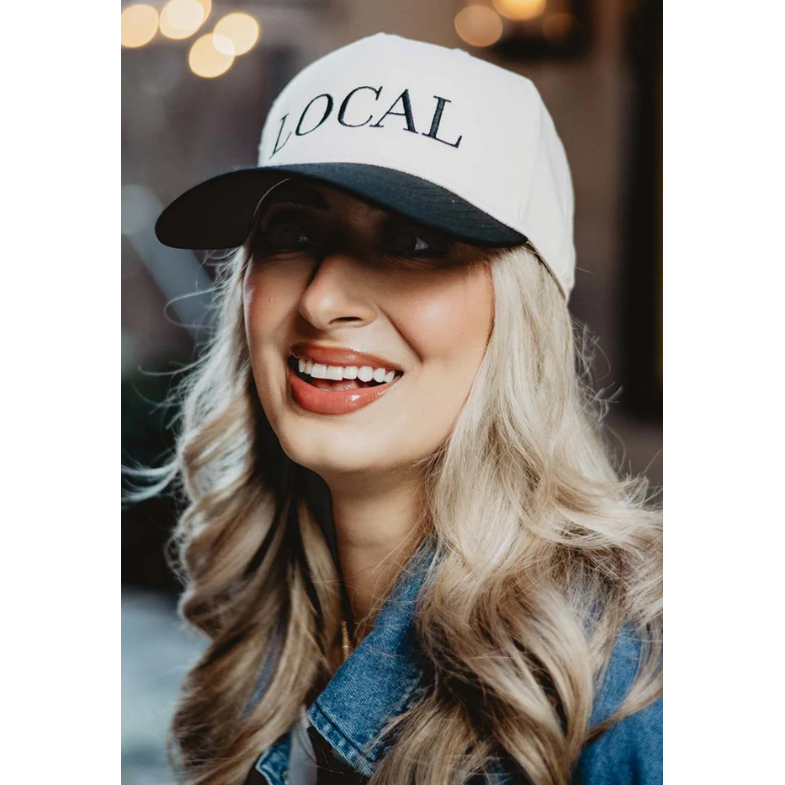 Photography of a smiling woman wearing a white and black "LOCAL" baseball cap, with long blonde wavy hair, glossy peach lips, and a denim jacket against a blurred background of warm bokeh lights