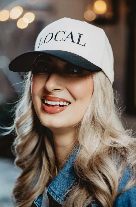 Photography of a smiling woman wearing a white and black "LOCAL" baseball cap, with long blonde wavy hair, glossy peach lips, and a denim jacket against a blurred background of warm bokeh lights