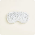 A fluffy light grey eye mask with a soft shaggy texture resting on a plain cream background featuring a small white care label on the right side