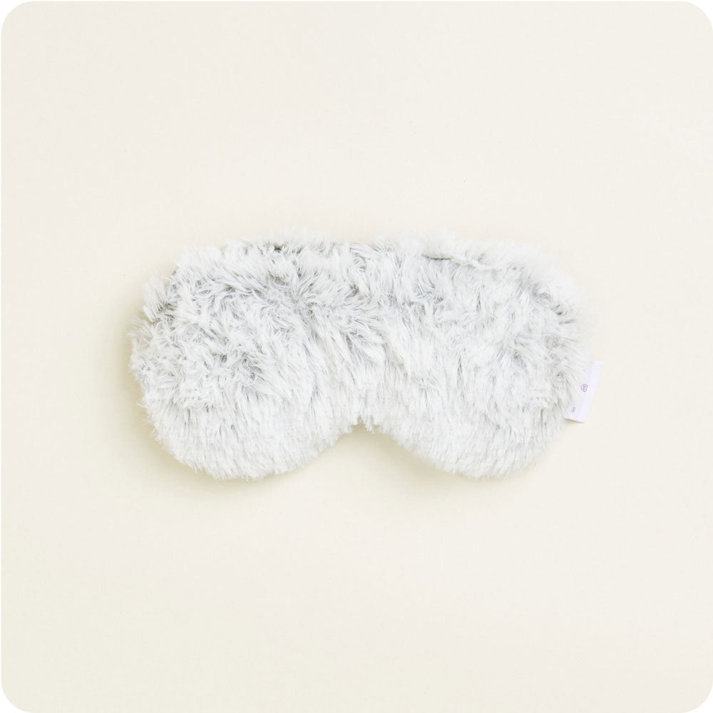 A fluffy light grey eye mask with a soft shaggy texture resting on a plain cream background featuring a small white care label on the right side