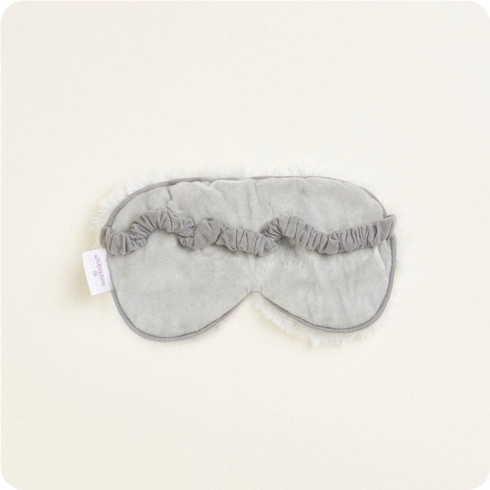 Product photo of a Warmies gray plush sleep mask on a light studio background featuring a ruffled elastic strap, white fluffy trim, and a visible Warmies brand tag