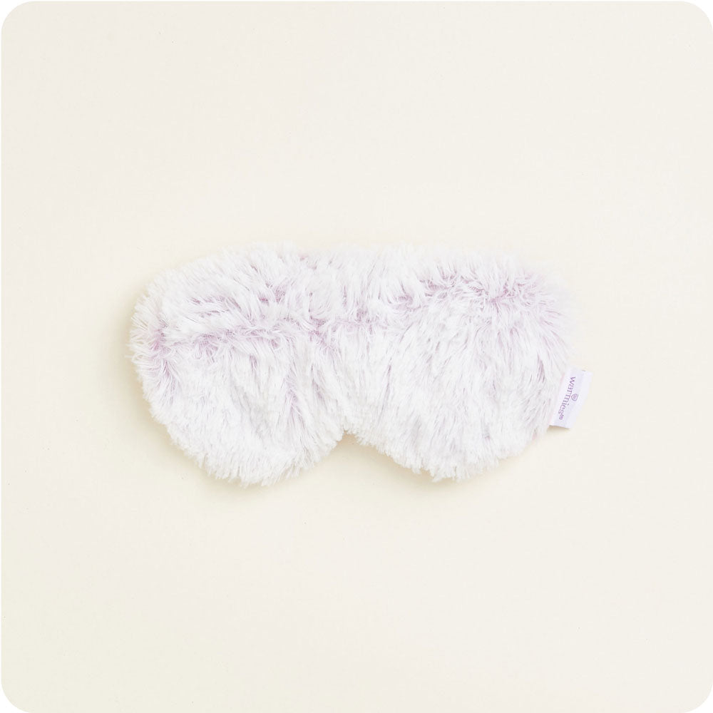 A light purple and white fluffy faux fur eye mask with a small "Warmies" brand tag visible on the right on a plain light background