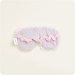 Product photograph of a soft lavender sleep mask with fuzzy white trim a ruffled pink elastic band and a Woollies brand tag laid flat on a cream background