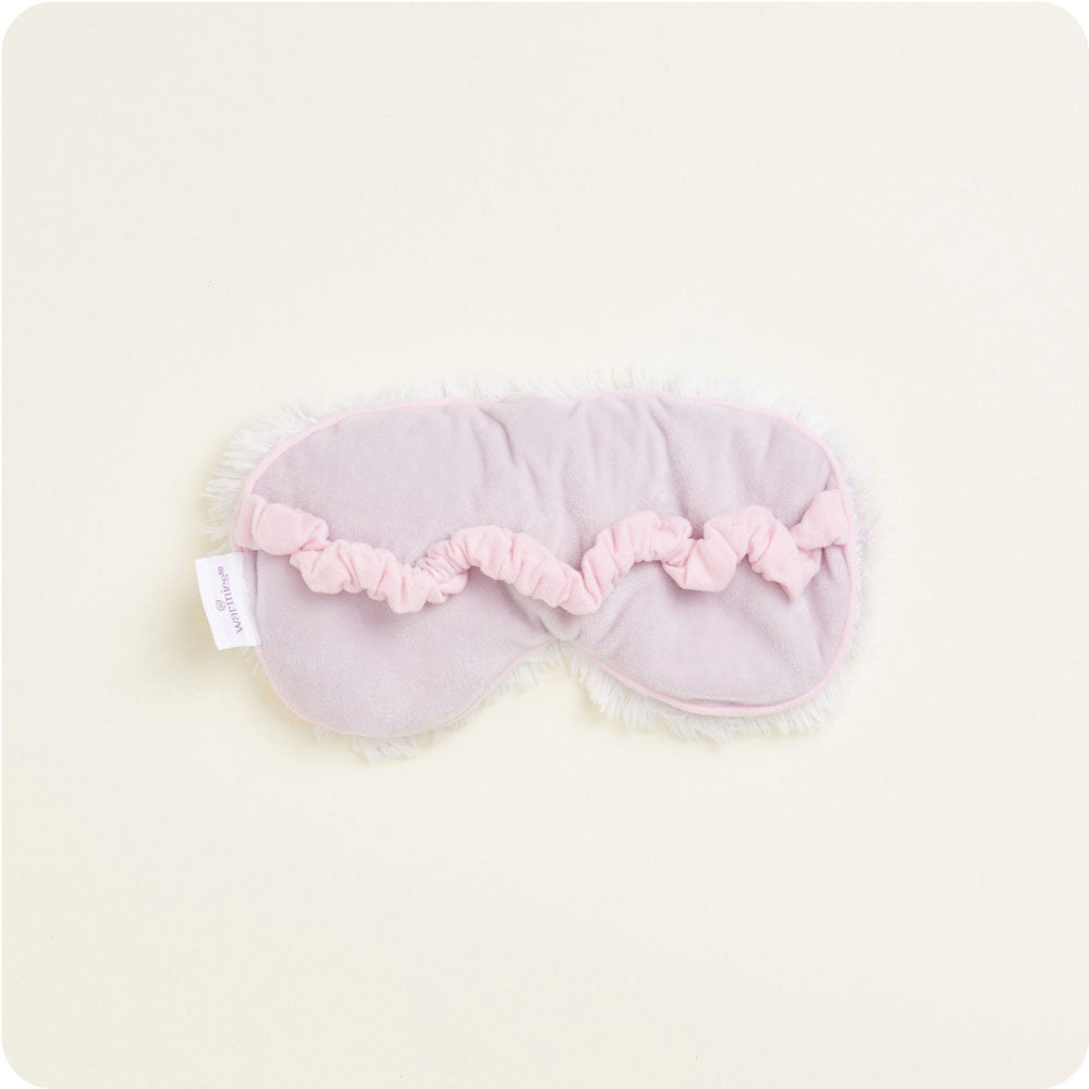 Product photograph of a soft lavender sleep mask with fuzzy white trim a ruffled pink elastic band and a Woollies brand tag laid flat on a cream background
