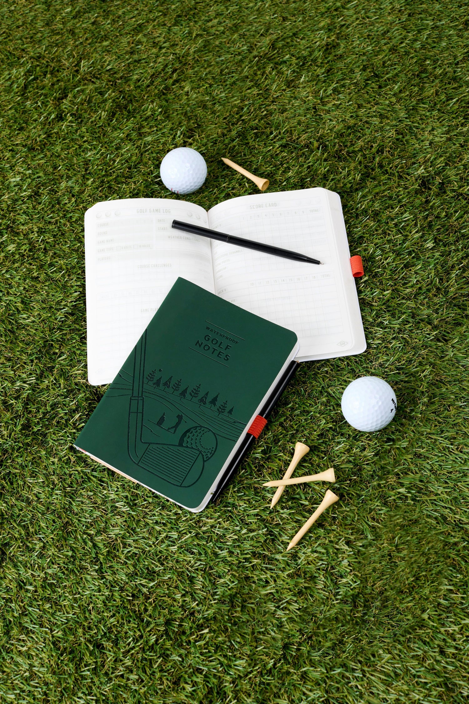 Flat lay of golf accessories on vibrant green artificial grass featuring a dark green "Waterproof Golf Notes" notebook with an embossed golf course design, an open "Golf Game Log" displaying score cards and a black pen with a red grip, two white golf balls, and three wooden tees