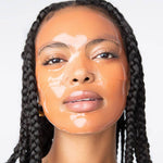 Photograph of a woman with dark skin and black braided hair wearing a translucent peach-toned sheet mask giving her face a shiny wet appearance with slightly parted lips