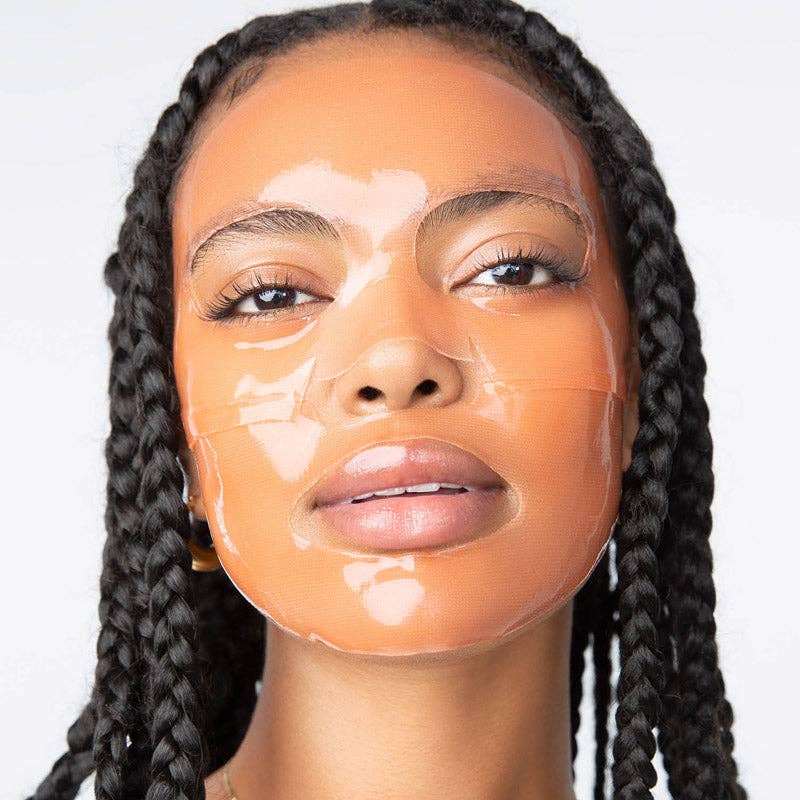 Photograph of a woman with dark skin and black braided hair wearing a translucent peach-toned sheet mask giving her face a shiny wet appearance with slightly parted lips