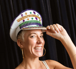 A smiling woman with short blonde hair wears a festive white captain's hat adorned with purple, green, and gold spikes, large gold hoop earrings, and a joyous expression against a dark pleated background