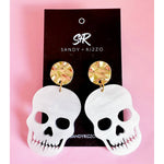 Sandy + Rizzo earrings featuring white matte skull drop earrings with detailed tooth cutouts and gold-toned hammered stud tops displaying abstract green pink and gold swirl patterns presented on a black card against a vibrant pink background