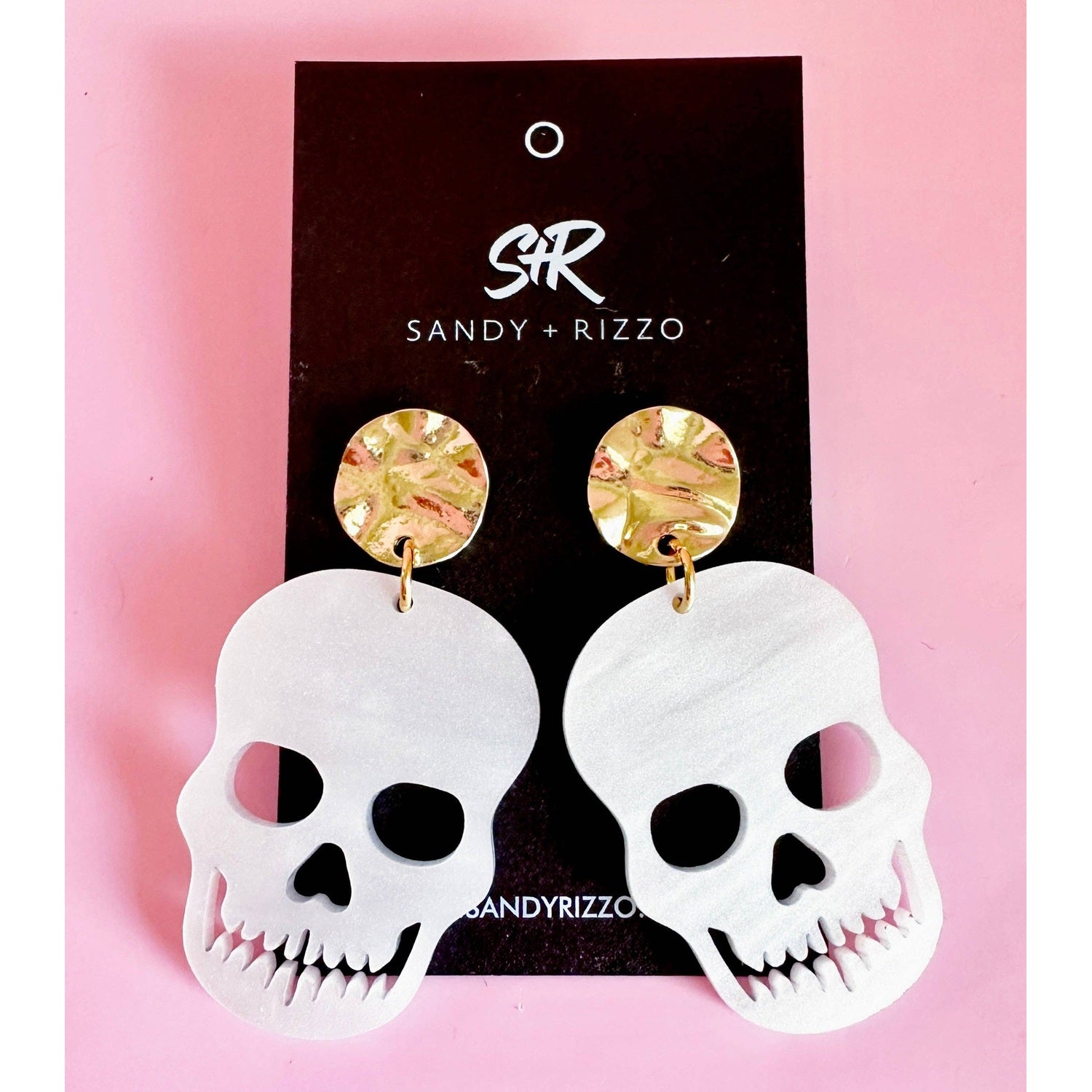 Sandy + Rizzo earrings featuring white matte skull drop earrings with detailed tooth cutouts and gold-toned hammered stud tops displaying abstract green pink and gold swirl patterns presented on a black card against a vibrant pink background