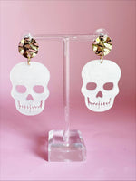 Acrylic white skull drop earrings with gold hammered stud tops and distinct jagged teeth cutouts presented on a transparent stand against a pastel pink background