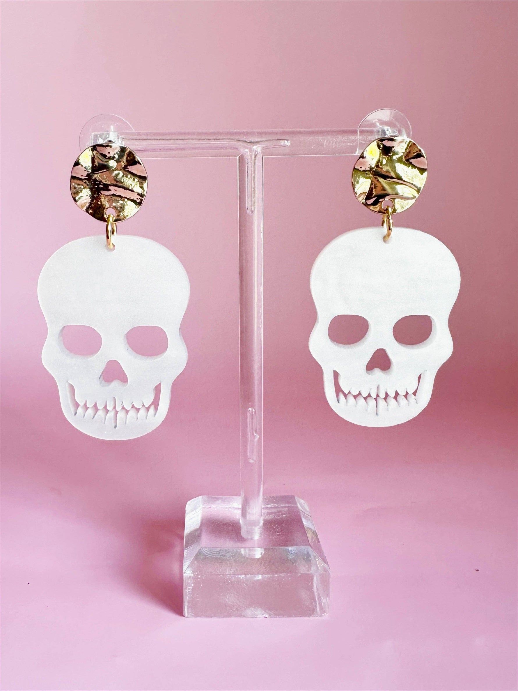 Acrylic white skull drop earrings with gold hammered stud tops and distinct jagged teeth cutouts presented on a transparent stand against a pastel pink background