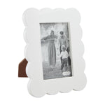 White scalloped photo frame with a brown easel back, displaying a black and white family portrait of a smiling mother, father, and toddler walking near a lake, featuring a small mud pie brand tag